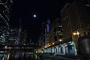 Fototapeta na wymiar Chicago city riverwalk promenade at night with vintage drawbridge, illuminated urban downtown buildings and the moon.