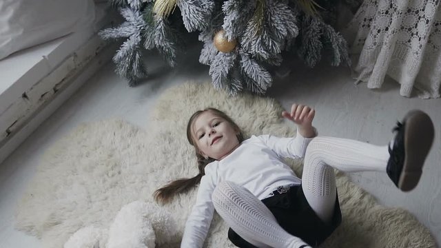 The charming girl lies on a soft white carpet and is played with a large plush bear. Winter holidays. Christmas tree
