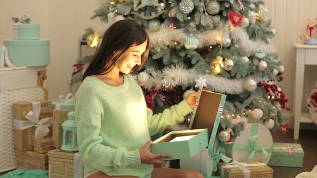 Happy pregnant girl opens a magical glowing Christmas gift while sitting near Christmas tree.