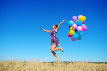 Beautiful, cheerful young woman in a bright dress with colorful balloons  on a field with a blue...