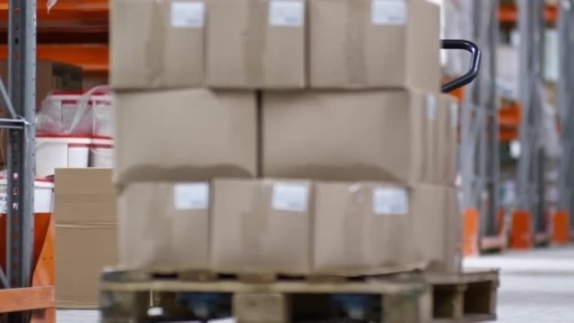 PAN of unrecognizable worker pulling cart with cardboard boxes through aisle of factory warehouse