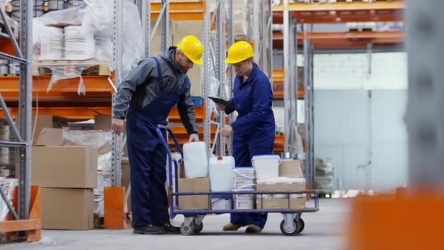 PAN of bearded male warehouse worker in hard hat carrying white jerrycan and putting it into platform cart, then talking with female colleague with tablet pointing and explaining something