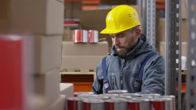 Zoom in with slowmo of bearded male worker in hard hat stocking cans of paint in warehouse and chatting on mobile phone