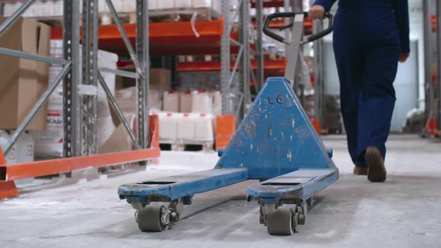 Tracking with low-section of unrecognizable female worker in blue overalls pulling empty platform cart through warehouse with rack shelves