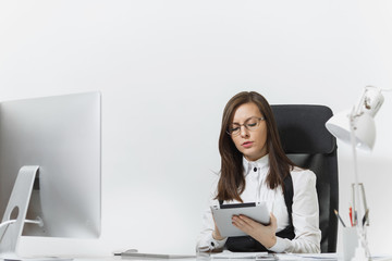 Beautiful serious engrossed brown-hair business woman in suit and glasses sitting at the desk,...