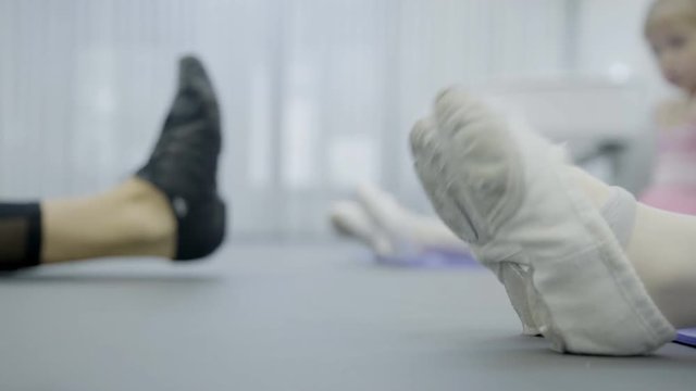 The prima ballerina teaches two girls how to point toes in the class. Little dancers sit on the right and wears white gym shoes and the teacher is on the left in the black footwear. The women stretch