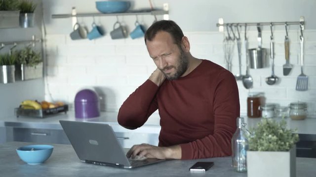 Young man having neck pain during work on laptop at home
