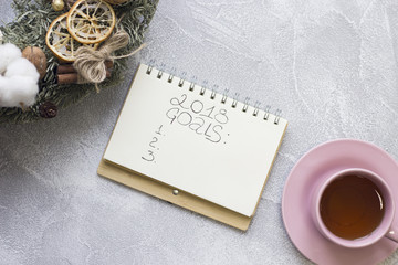 2018 goals inscription in a notebook, pink tea cup with saucer, christmas wreath with natural decorations on a light concrete background, top view