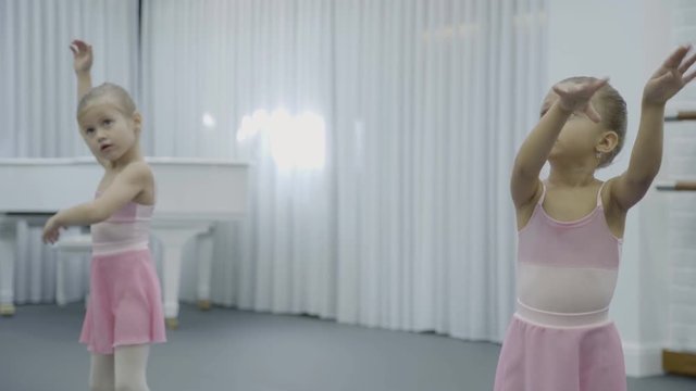 Two girls do the trunk turnings with rasing hands during the ballet class. Little dancers in pink bresses and with buns stand in the big bright room and exercising. The white silk curtain and the