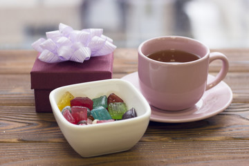 A festive still life with a pink tea cup with saucer, a bowl of colored candies and jelly beans and a purple gift box with a light purple bow on a wooden table