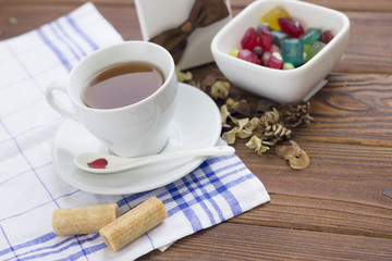 Still life with a white porcelian tea cup,saucer and spoon on a white blue plaid napkin,a bowl of colored candies and a white gift box with a brown bow on a wooden table