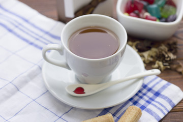 Still life with a white porcelian tea cup,saucer and spoon on a white blue plaid napkin and a bowl of colored candies on a wooden table