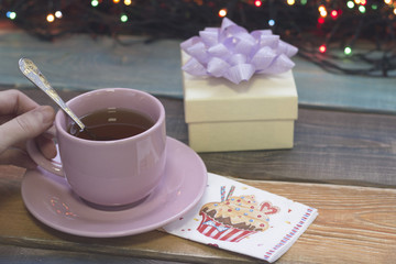 Festive still life with a pink cup of tea,saucer and spoon, a light gift box with a purple bow,a decorated napkin and fairy lights, colored wooden background