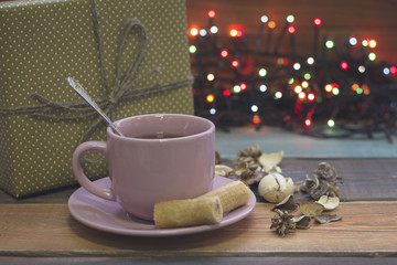Obraz na płótnie Canvas Festive still life with a pink porcelian cup of tea,a saucer, a teaspoon, cookies, a golden giftbox and fairy lights, colored wooden background