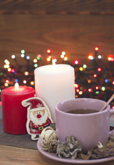 Festive still life with a pink porcelian cup of tea, a saucer, a teaspoon, toy santa, two burning candles and fairylights on the back, a colored wooden background