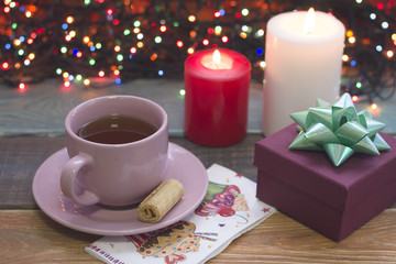 Obraz na płótnie Canvas A festive still life a pink porcelian cup of tea, a saucer, a purple gift box with a shiny green bow, a decorated napkin, two burning candles and fairylights on the back, a colored wooden background