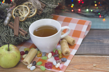 A festive still life with a cup of tea on a plaid orange napkin, a fresh green apple, spilled colored candies candies and a pine wreath on a colored wooden table