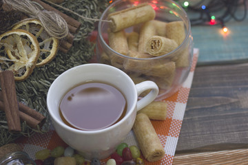 A festive still life with a cup of tea on a plaid orange napkin, a a bowl of cookies and a pine wreath on a colored wooden table