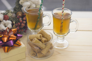 A bright festive morning with two clear cups of greentea, bowl of cookies and a gift box with a shiny bow, Christmas pine wreath on a wooden table, light background