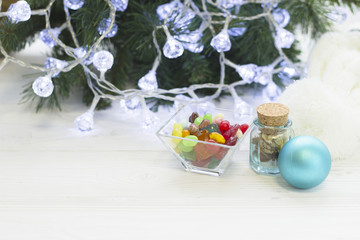 Fototapeta na wymiar A square clear bowl of colored candies, a tiny jar of dry flowers and a Christmmas-tree blue ball by a Cristmas-tree decorated with white fairy lights, light wooden background