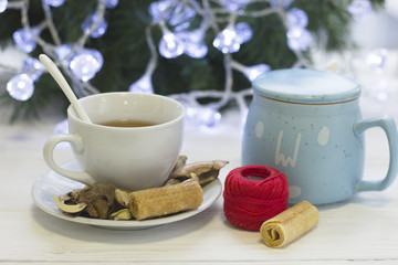 A festive bright morning with a white porcelian cup of hot black tea with a spoon, a light blue sugar bowl, dry flowers, cookies and a skein of red threads, blurred Christmas tree with white fairy