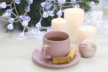 Obraz na płótnie Canvas A festive still life with a pink tea cup, a saucer, a pink shiny Christmas-tree ball, three white burning candles, a blurred Christmas tree with fairy lights on the back, light background