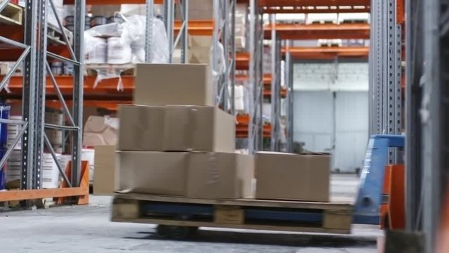 PAN of unrecognizable male worker in blue overalls pulling cart with cardboard boxes through factory warehouse with rack shelves