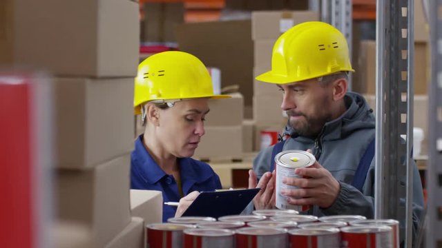 Zoom in of male bearded worker in hard hat standing before rack shelves and inspecting cans of paint while female colleague in uniform writing on clipboard and talking
