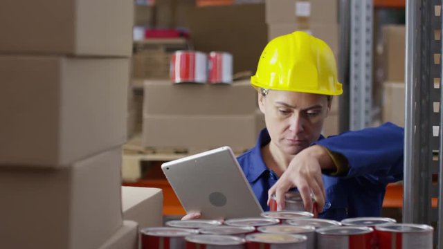 Zoom out with slowmo of concentrated female warehouse worker wearing uniform and hard hat standing before rack shelf and inspecting cans of paint, then typing on tablet