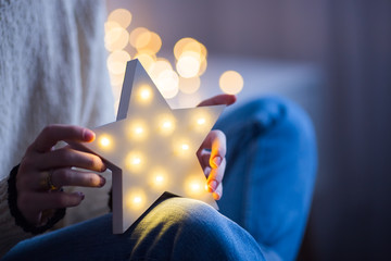 Young female hands holding glowing white LED star on bokeh background indoor