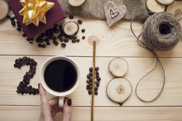 Fototapeta na wymiar 2018 inscription of coffee beans, woman's fingers holding a mug and wooden slices with a purple gift box and skein of threads on light wooden background, top view