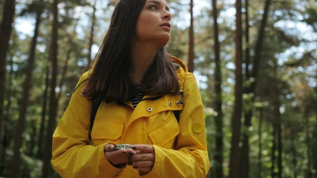 Young beautiful worried woman in yellow coat and backpack looking for the way back with a help of a compass in the middle of the forest. Got lost. Outdoors