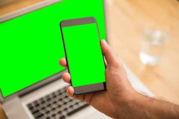 Young adult man hand hold green screen smartphone in front of green screen computer in bright office in the morning.