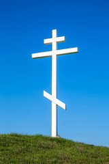 A wooden cross on top of a hill. Orthodox white cross glows on the top of the hill on the background of blue sky and of green grass