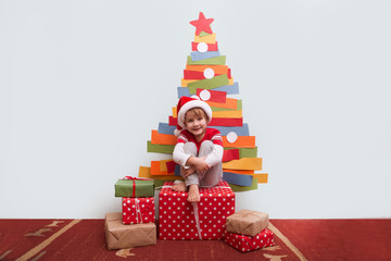 Children boy in Santa red hat  with decoration for Christmas and Christmas present. Christmas time. paper Christmas tree on a wall in the room.