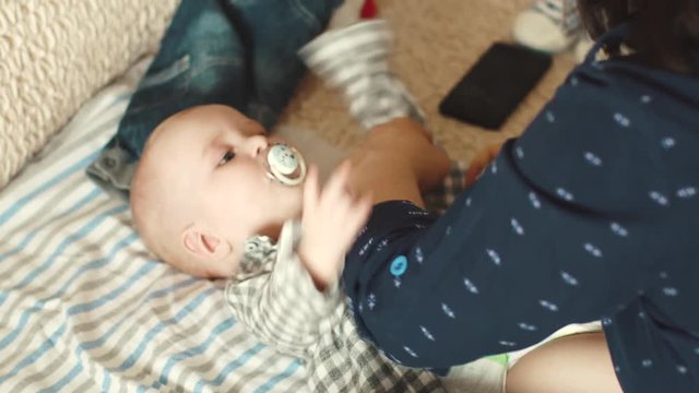 Mom puts on a shirt on her little son. mother takes care of the child. baby holds a pacifier in his mouth