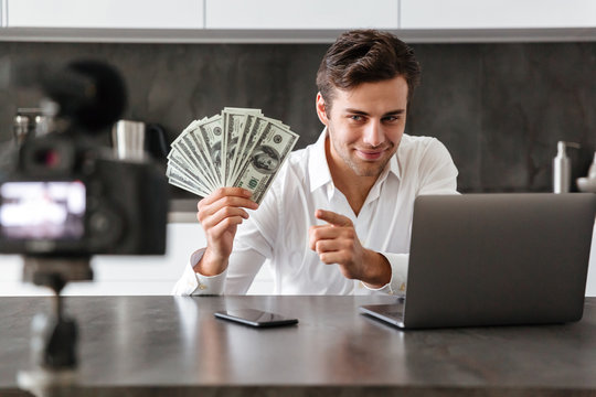 Smiling young man filming his video blog episode about new tech devices while sitting at the kitchen table with laptop and showing bunch of money banknotes