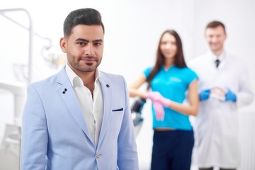 Handsome young man smiling to the camera posing at the dental clinic his dentist and nurse standing on the background copyspace professionalism trust happiness service dentistry oral smile.
