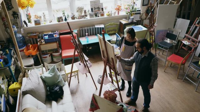 Skilled artist man teaching young woman painting on easel at art school studio - creativity, education and art people concept