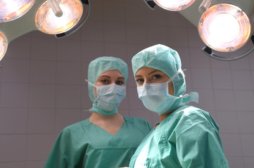 Two young women pose in a low lit operation theater.  Fully dressed as theater nurses with face masks  and green sterile medical work clothing.