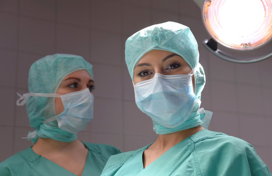 Two young women pose in a low lit operation theater.  Fully dressed as theater nurses with face masks  and green sterile medical work clothing.