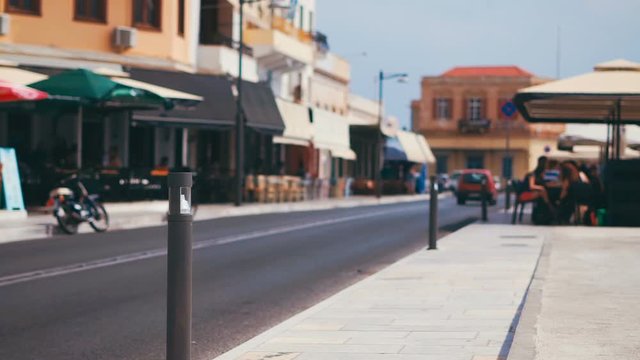 Defocused cars drive along the tourist street with restaurants in mediterranean European historic resort town at bright sunny day. Ermoupolis, Syros island, Greece.