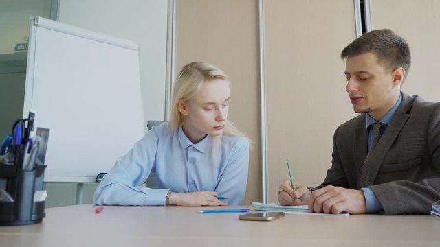 The businessman making remarks in the report of his young female subordinate in his private office. He writes by a pencil on the papers laying in front of him and the lady in the table. The boss wears
