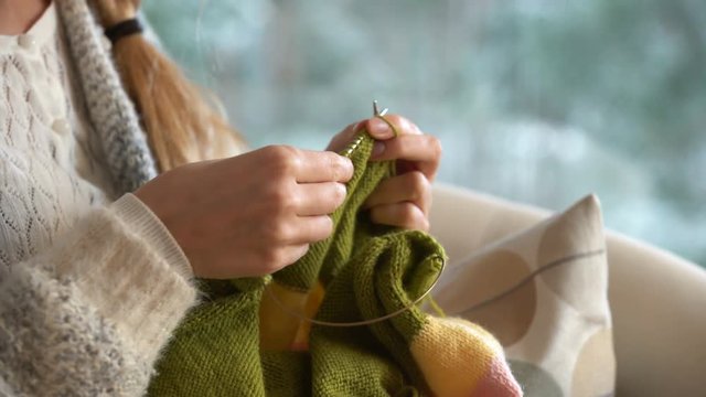 Cozy winter lifestyle. Young woman knitting warm wool sweater in the sitting room against snow landscape from outside.