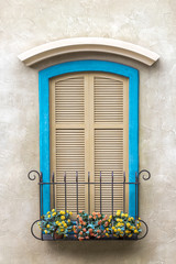 Two windows, with bright blue frames, and shutters on the glass. The windows have flowers at the base. A quaint scene.