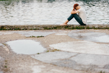 Barefoot lonely unknown adult man sitting on edge of embankment outdoor. Homeless poor person in depression. Waiting for help. Problematic life. Psychological dramatic male portrait. Friendless person