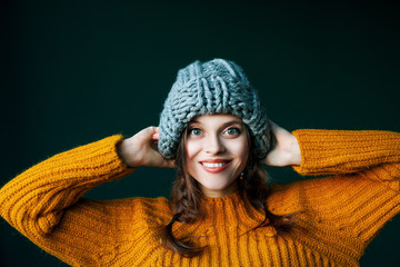 Close up portrait of young beautiful happy smiling girl wearing stylish grey big loop knitted beanie hat and yellow sweater. Model looking at camera, posing on dark green background.