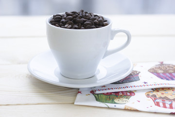 Christmas still life with white coffee cup with saucer on a wooden table. The cup filled with coffee beans. Light background. Close up