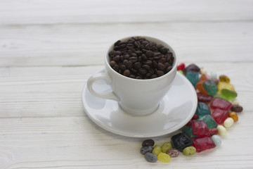 Festive still life with a cup of coffee with a saucer and spilled colored candies. The cup is filled with coffee beans. Close up. Light wooden background. Copy space
