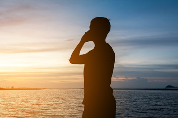 Silhouette of man standing alone on tropical beach with calm blue sea and holding smartphone.